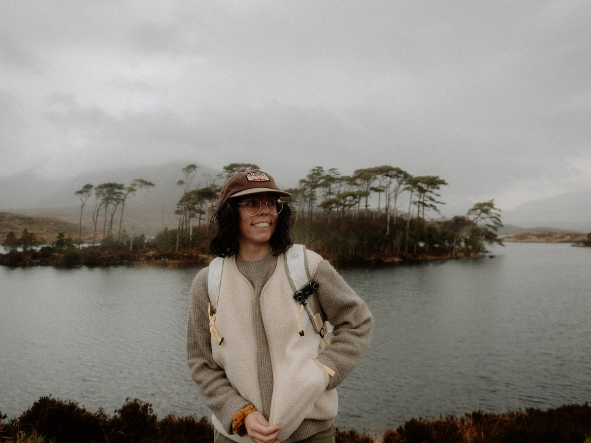 A woman wearing a beige fleece vest over a gray sweater, a brown cap, and a backpack stands in front of a misty lake and small island forest under an overcast sky.