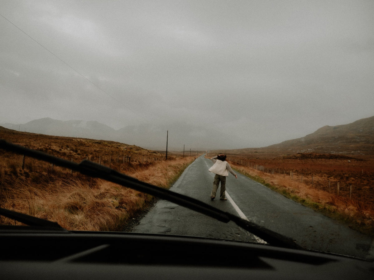 A woman walking down the middle of a deserted road, wearing a beige fleece, with dramatic landscape all around. Photo taken from inside a vehicle