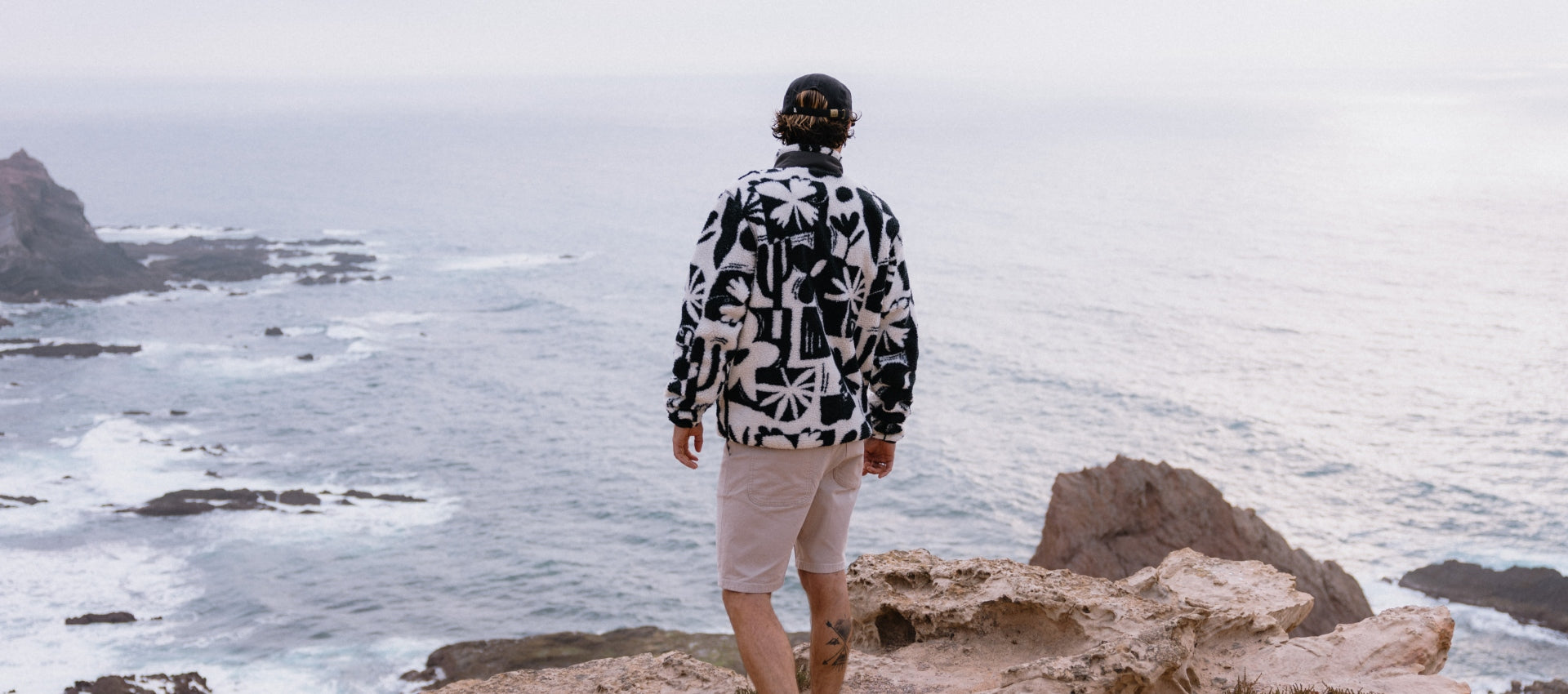 A man looking out over the sea, standing on a cliff, wearing a black and white patterned fleece