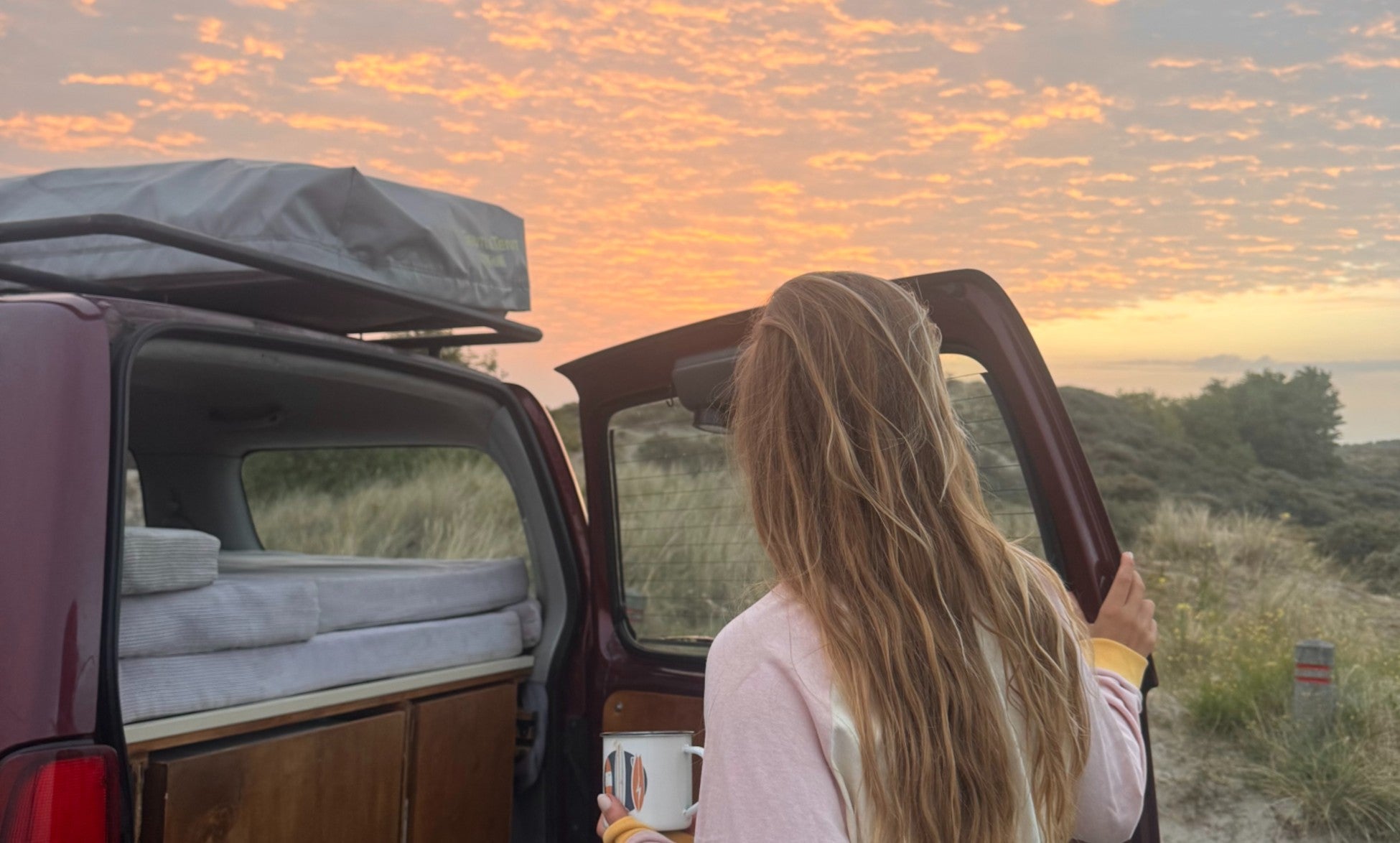 A woman looking out at a sunset sky, standing next to the open back door of her suzuki jimny
