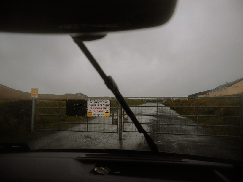 View from inside a vehicle looking out at a wet gate and road under an overcast sky, with a sign reading 'Access to the cliffs is closed'
