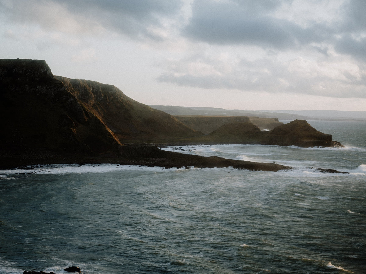 A scenic coastal landscape with dark rocky cliffs rising from a blue-gray ocean under a cloudy sky.