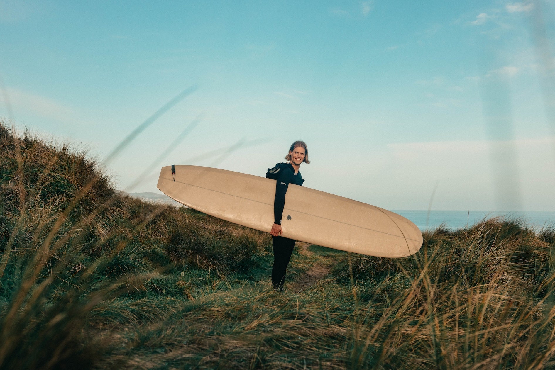 Person holding a surfboard on a grassy hill with a clear sky